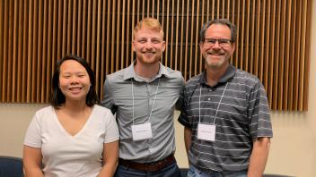 Three individuals smiling indoors in front of wooden slatted wall panels at what appears to be a professional or academic event.