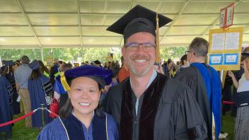  Two people in academic regalia posing together at an outdoor UC Davis graduation ceremony under a large tent.
