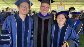 Three individuals in academic regalia smile together under a large canopy at a graduation ceremony.