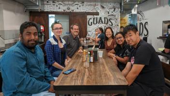 A group of seven people sit around a wooden table at a casual restaurant, smiling and enjoying a meal together.