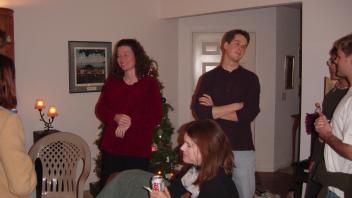 People standing and chatting near a Christmas tree in a warmly lit living room.