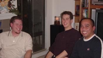 Three men sitting and smiling in a living room with a bookshelf and TV in the background.