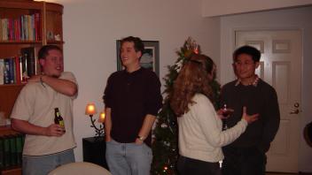 Group chatting and laughing at a holiday gathering, with bookshelves and a Christmas tree visible.