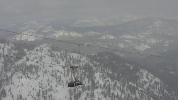 A tram car suspended mid-air travels above snowy pine-covered mountains under a gray, overcast sky.