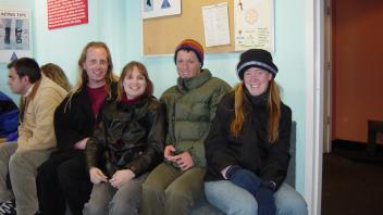 Four people sitting indoors on a bench in winter gear, smiling at the camera near a ski resort bulletin board.