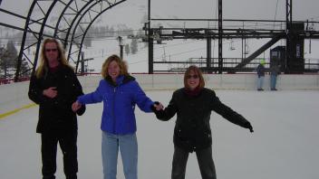 Three people holding hands while ice skating on an outdoor rink, all wearing coats and smiling as snow falls around them.