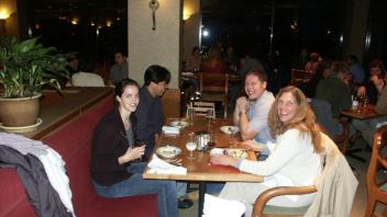 Group of four people enjoying dinner at a restaurant table, smiling and engaged in conversation with others in the background.