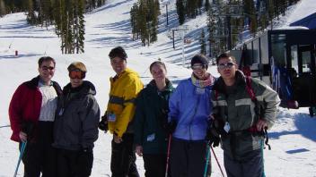Group of six skiers posing at the base of a snowy slope with ski lifts and pine trees in the background.