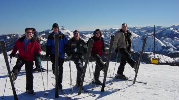 Five skiers standing with skis planted in the snow at a mountain summit with panoramic alpine views.