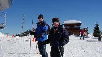 Two skiers in jackets and sunglasses pose near a ski lift on a sunny snow-covered mountain.