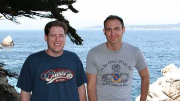 Two men standing on a scenic ocean cliff with trees and distant shoreline in the background.
