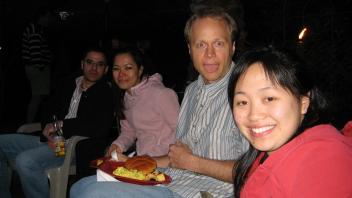 Four people sitting outside at night, smiling at the camera with food and drinks in hand.