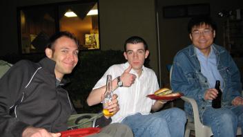 Three men sitting on lawn chairs at night, holding drinks and food, smiling and giving a thumbs up.