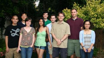 Group photo of nine people standing in a backyard with greenery in the background.