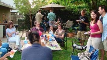  Outdoor gathering with people sitting and standing around tables enjoying food.
