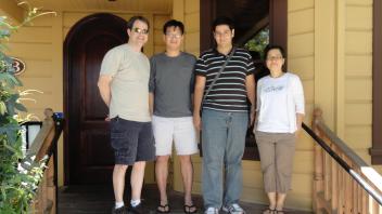 Four people posing on the porch of a wooden house with a rounded door.