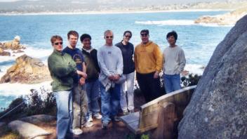 Eight people posing along a coastal trail with ocean and cliffs in the background.
