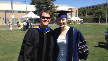 Faculty and graduate in full academic regalia pose together outdoors on graduation day.