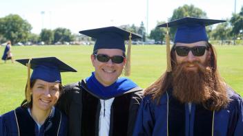 Three graduates in caps and gowns smiling outdoors on a sunny day.