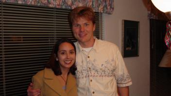  A woman and a man pose together inside a house near a window with floral curtains.