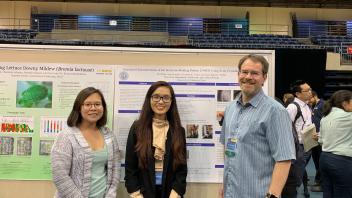 Two students and a faculty member smile for a photo by the L194D1 poster.