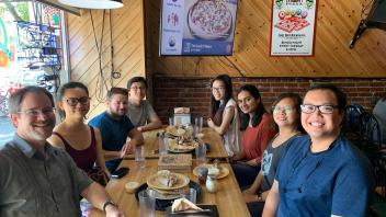 A group of students and faculty enjoy a casual group meal at a wooden table in a restaurant.