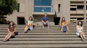 Five individuals sitting spaced apart on concrete steps in front of a beige academic building, with graduation sashes on two of them.