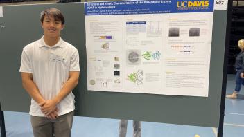 Student standing alone beside a UC Davis research poster presentation on RNA-editing enzyme ADAR, inside a gymnasium.
