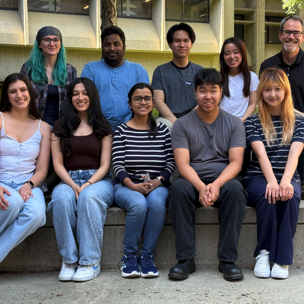 A group of ten lab members pose for a group photo on outdoor concrete steps in front of a campus building, smiling together in casual attire on a sunny day.